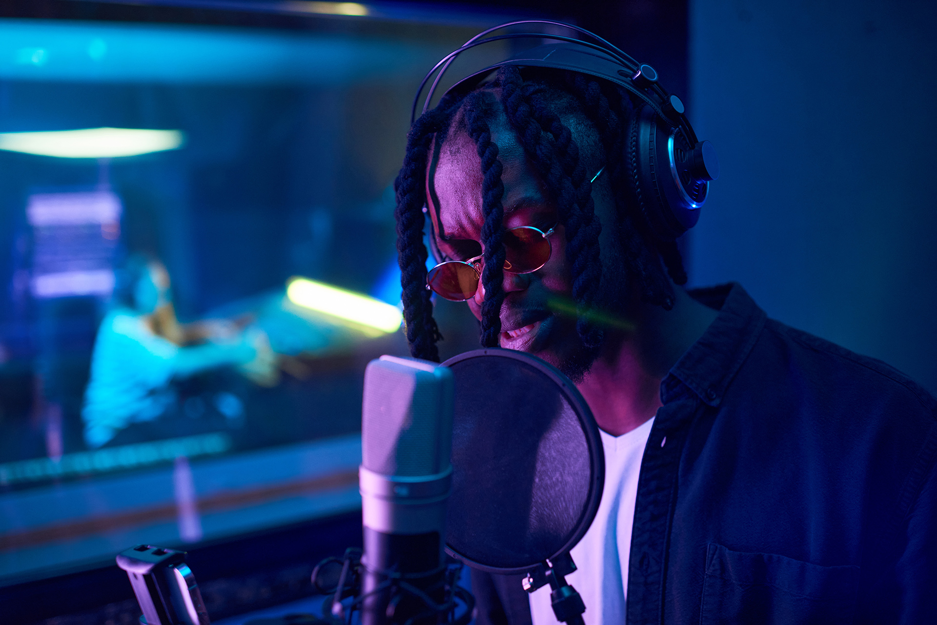 African young man in sunglasses with dreadlocks wearing headphones and singing to microphone his song in the studio
